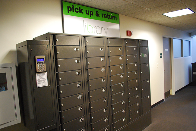 Dark grey lockers with a large sign overhead that reads Pick Up & Return, with library logo underneath