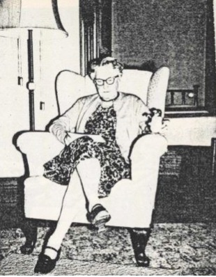 Black and white photo of woman with short hair and glasses, sitting in a light colored wingback chair, reading