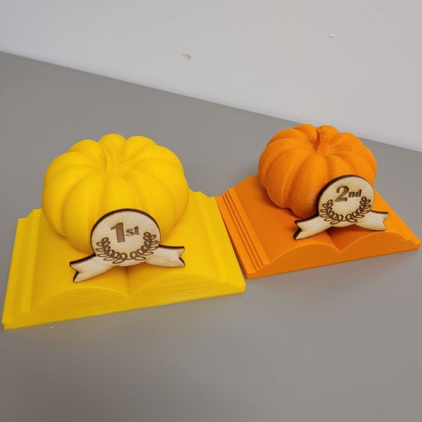 Pumpkin trophies printed in yellow and orange filament. First and second place ribbons etched and cut out of wood on the laser cutter, glued to the pumpkin with tacky glue.
