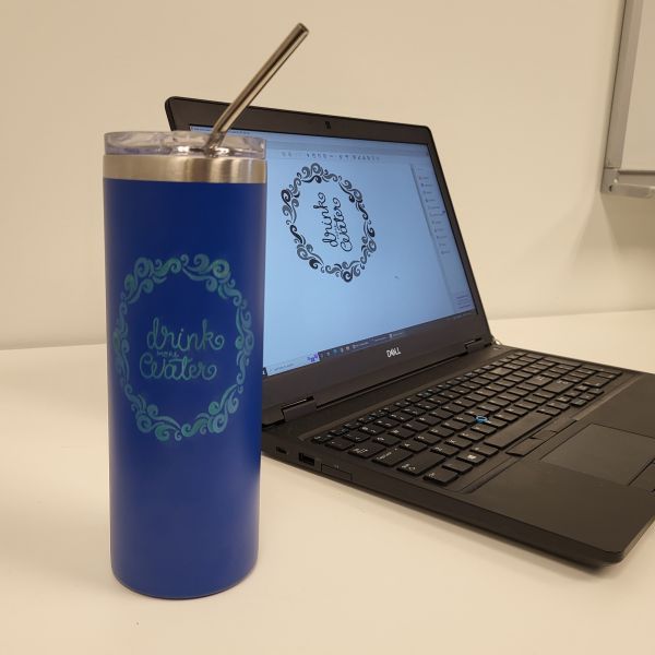 Blue powdercoated drinking tumbler etched with a flowy frame and the words Drink More Water. The file sent to the laser can be seen on the computer screen behind the tumbler.