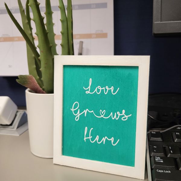 White frame and teal back panel glued together to make a frame. Love Grows Here cut out of white adhesive vinyl is placed onto the teal background.