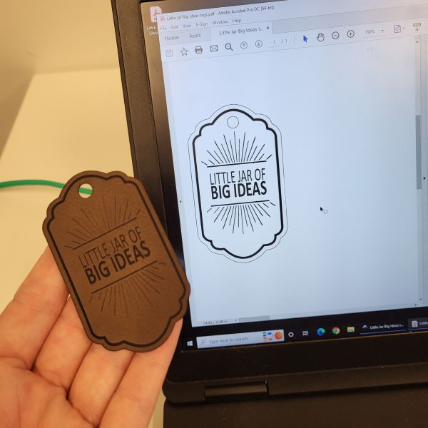 Dark brown leather-like tag that says Little Jar of Big ideas. Design file sent to the laser can be seen on the computer screen.