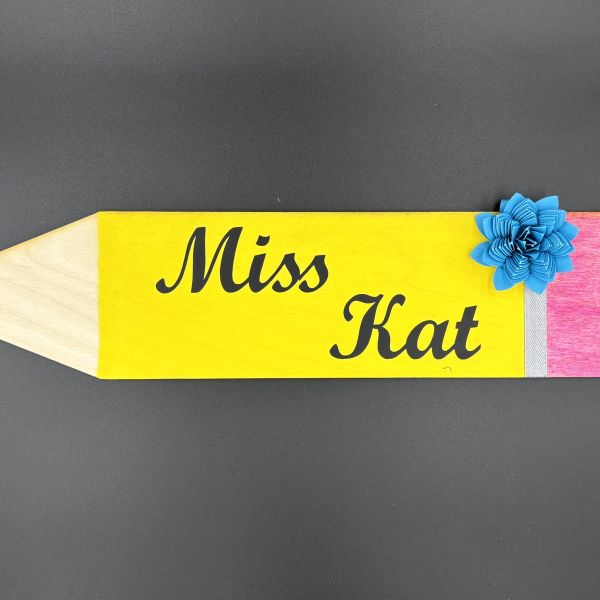 Pencil shape cut out of wood and painted with acrylic paint. Blue paper flower embellishment and the words Miss Kat cut out of black vinyl and placed in the middle.