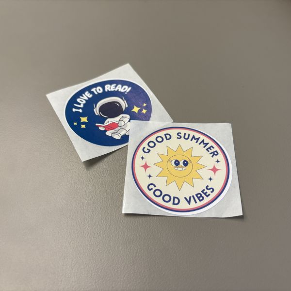Two round 2 inch stickers with cartoon-style images, one with a sun that says good summer good vibes, and the other is an astronaut reading that says I love to read.