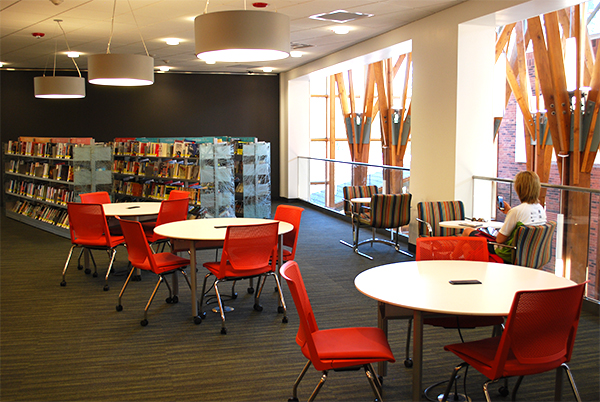 Photograph of library second floor balcony area, with round tables, red chairs, customer reading at one smaller table, bookshelves in the background